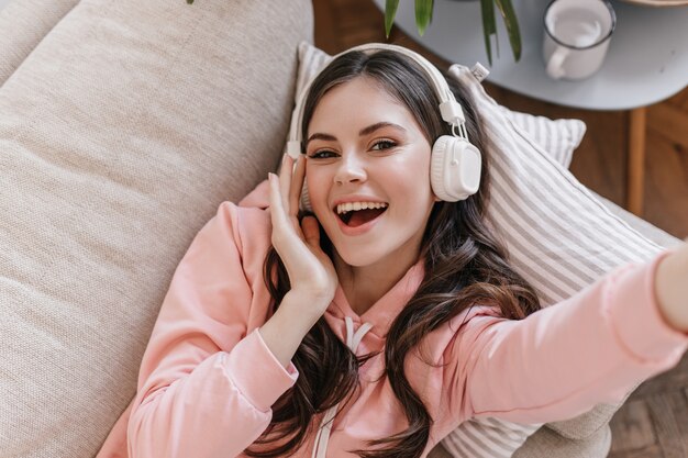 Mujer atractiva en sudadera rosa está acostada en el sofá, disfrutando de la música en auriculares y haciendo selfie