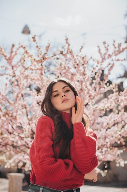 Mujer atractiva en suéter brillante mira a cámara sobre fondo de sakura. Instantánea de dama en suéter rojo posando afuera y disfrutando de la primavera