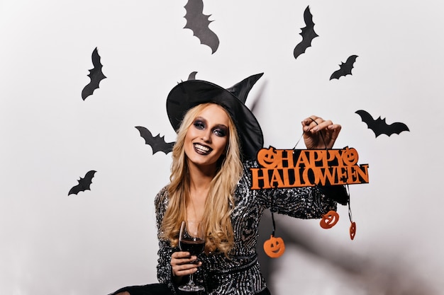 Mujer atractiva en traje de mago celebrando halloween. Impresionante chica rubia con sombrero de bruja disfrutando de la mascarada.