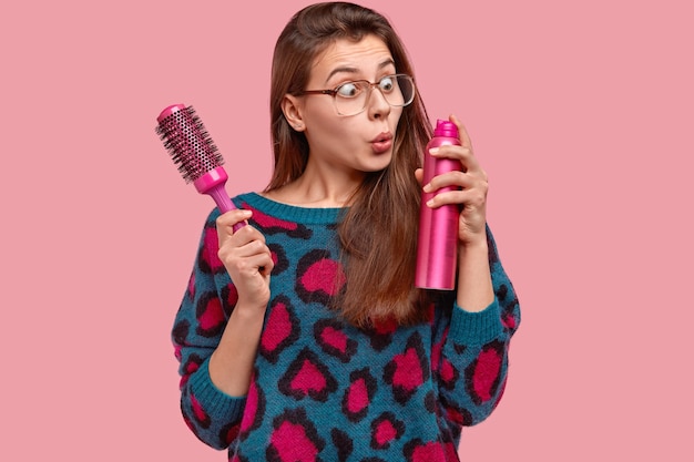 Foto gratuita mujer atractiva usa grandes gafas cuadradas, mira con estupefacción la laca para el cabello, sostiene el cepillo, quiere hacer un peinado para una ocasión especial