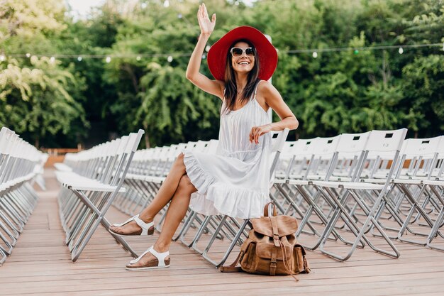 Mujer atractiva vestida con vestido blanco, sombrero rojo, gafas de sol sentado en el teatro al aire libre de verano en una silla sola, tendencia de moda de estilo callejero de primavera, accesorios, viajar con mochila, agitando la mano