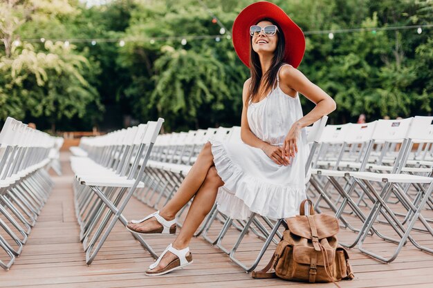 Mujer atractiva vestida con vestido blanco, sombrero rojo, gafas de sol sentado en el teatro al aire libre de verano en una silla sola, tendencia de moda de estilo callejero de primavera, accesorios, viajar con mochila