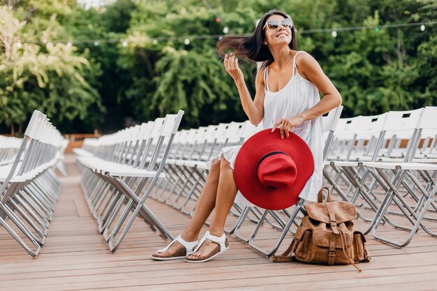 Mujer atractiva vestida con vestido blanco, sombrero rojo, gafas de sol sentado en el teatro al aire libre de verano en una silla sola, tendencia de moda de estilo callejero de primavera, accesorios, viajar con mochila
