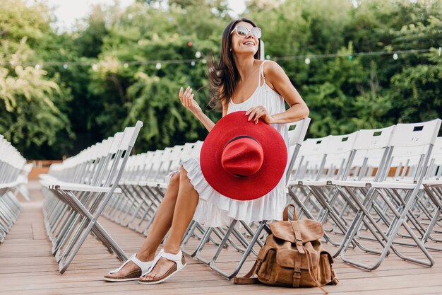 Mujer atractiva vestida con vestido blanco, sombrero rojo, gafas de sol sentado en el teatro al aire libre de verano en una silla sola, tendencia de moda de estilo callejero de primavera, accesorios, viajar con mochila