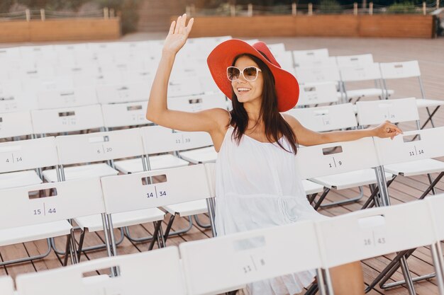 Mujer atractiva vestida con vestido blanco, sombrero rojo, gafas de sol sentado en el teatro al aire libre de verano en una silla sola, tendencia de moda de estilo callejero de primavera, distanciamiento social