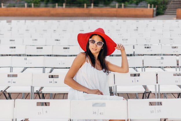 Mujer atractiva vestida con vestido blanco, sombrero rojo, gafas de sol sentado en el teatro al aire libre de verano en una silla sola, tendencia de moda de estilo callejero de primavera, distanciamiento social