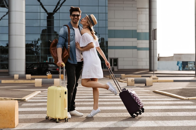Mujer atractiva con vestido blanco y sombrero besa a un hombre moreno guapo Los turistas geniales posa con maletas cerca del aeropuerto