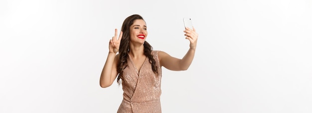 Mujer atractiva en vestido de lujo que muestra el signo de la paz en la cámara del teléfono tomando selfie en la posición del teléfono inteligente