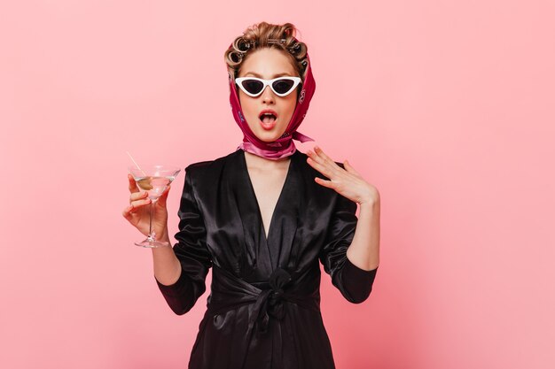 Mujer atractiva en vestido negro y pañuelo en la cabeza posando en la pared rosa con copa de martini