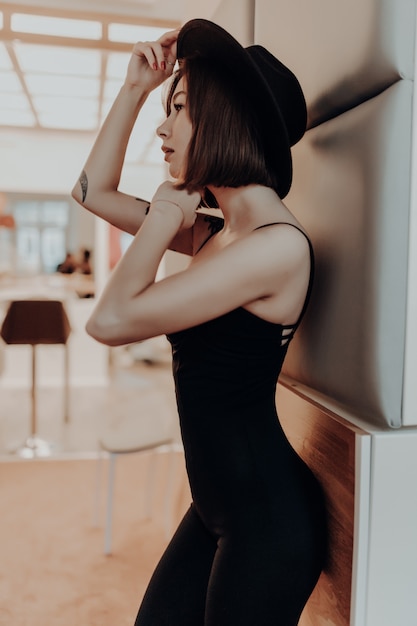 Mujer atractiva en vestido negro y sombrero de pie cerca de la pared en un apartamento de lujo