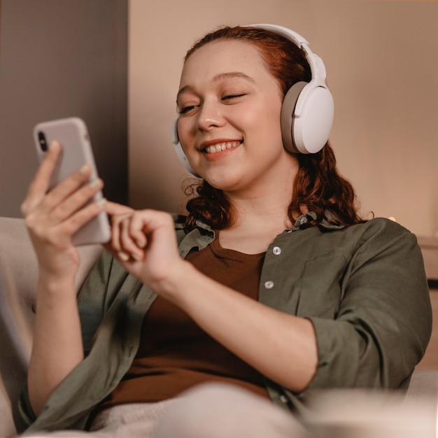 Mujer con auriculares modernos y dispositivo de teléfono inteligente en el sofá en casa