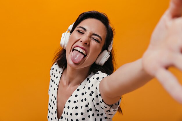 Mujer con auriculares muestra lengua y hace selfie sobre fondo naranja. Chica alegre se entrega a una blusa de lunares blancos.