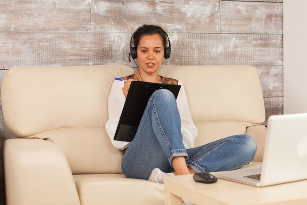 Mujer autónoma con auriculares y tomando notas en el portapapeles durante la videollamada de trabajo.
