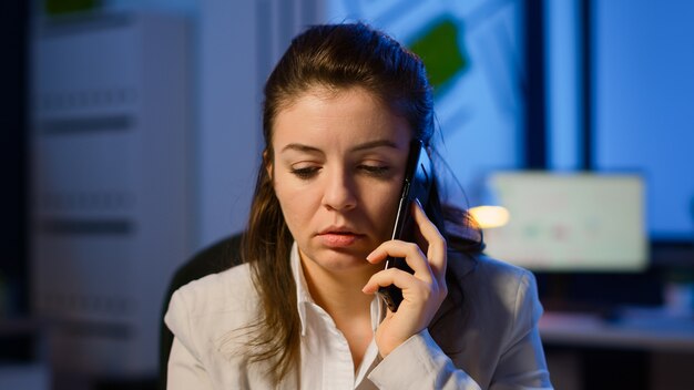 Mujer autónoma cansada que tiene conversación por teléfono mientras trabaja agotado en la oficina de negocios a altas horas de la noche haciendo horas extraordinarias. Empleado centrado en el uso excesivo de trabajo inalámbrico de la red de tecnología moderna