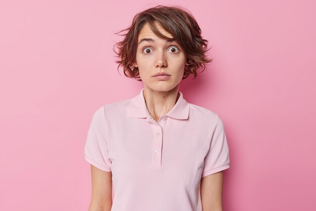 Mujer avergonzada conmocionada con peinado corto mira fijamente los ojos saltones se siente ansiosa vestida con una camiseta casual aislada sobre fondo rosa parece incrédula escucha algo impresionante. reacciones humanas