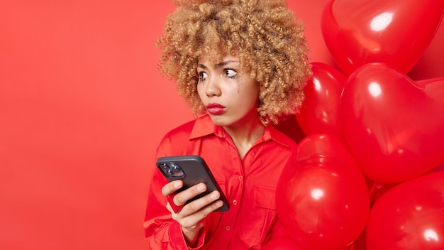 Una mujer avergonzada y sorprendida mira hacia otro lado con una expresión atónita y ve algo asombroso e impresionante que usa el teléfono inteligente para poses de comunicación en línea con un montón de globos contra la pared roja del estudio