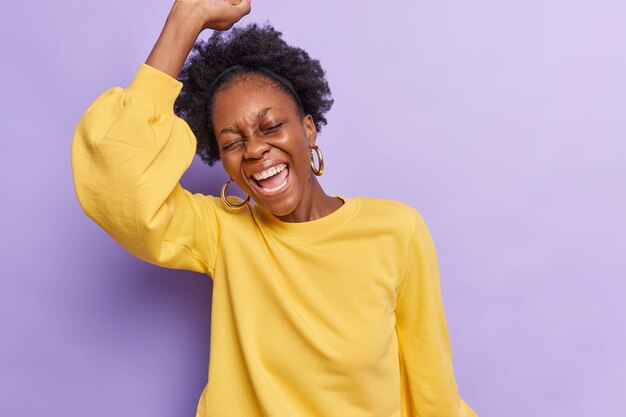 Mujer baila con alegría mantiene el brazo levantado exclama alegremente celebra algo vestido con un jersey amarillo casual aislado en violeta