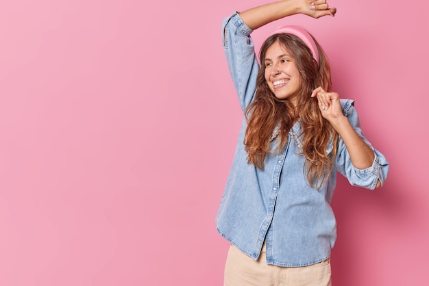 mujer baila con los brazos levantados sonríe ampliamente se mueve activamente usa diadema y camisa de mezclilla aislada en el espacio de copia rosa a la izquierda para su contenido promocional