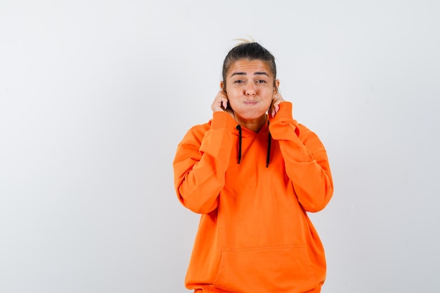 Mujer bajándose los lóbulos de las orejas en una sudadera con capucha naranja y mirando gracioso