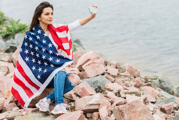 Mujer con bandera americana tomando selfie al lado del agua