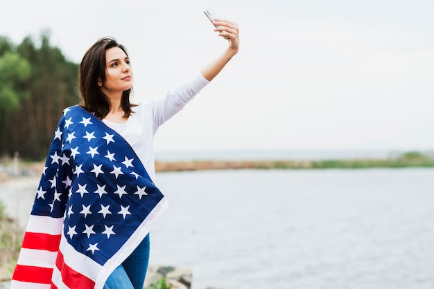 Mujer con bandera americana tomando selfie por un lago