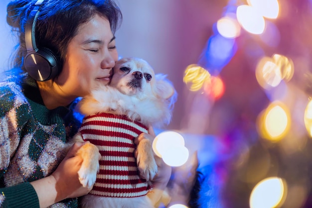 Una mujer bastante asiática escucha música de un auricular, un abrazo de mano, un lindo amigo perrito faldero con cuidado y amor, ternura, un momento alegre, una mujer asiática sostiene una pequeña mascota en el sofá con un árbol de decoración de Navidad.