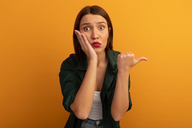 Mujer bastante caucásica ansiosa pone la mano en la cara y señala al lado en naranja