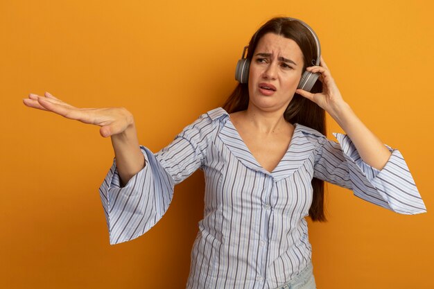 Mujer bastante caucásica disgustada en auriculares sostiene la mano abierta y mira al lado en naranja