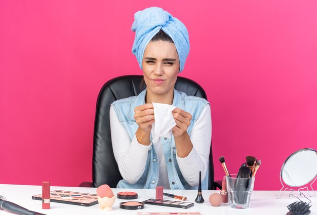 Mujer bastante caucásica disgustada con el pelo envuelto en una toalla sentada en la mesa con herramientas de maquillaje sosteniendo una servilleta de inserción aislada en la pared rosa con espacio de copia