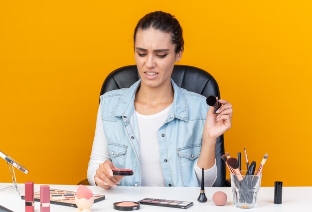 Mujer bastante caucásica disgustada sentada a la mesa con herramientas de maquillaje sosteniendo pincel de maquillaje y mirando rubor aislado en pared naranja con espacio de copia