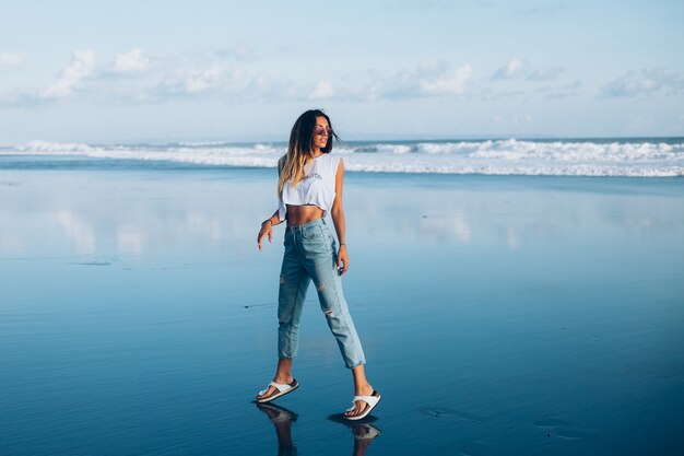 Mujer bastante caucásica en forma en top blanco y jeans en la playa reflectante por el océano a la luz del atardecer