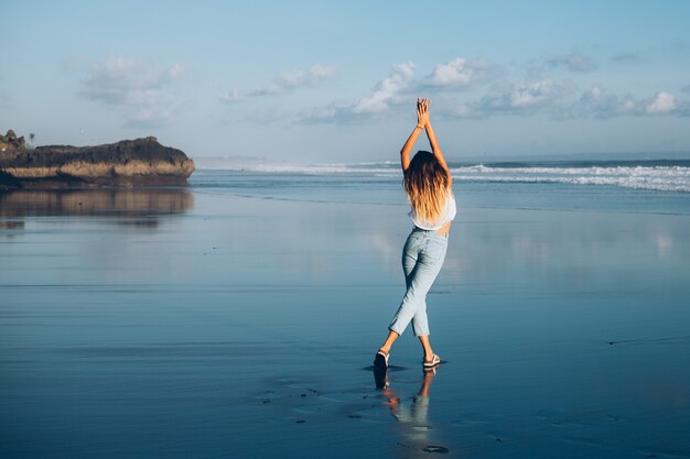 Mujer bastante caucásica en forma en top blanco y jeans en la playa reflectante por el océano a la luz del atardecer