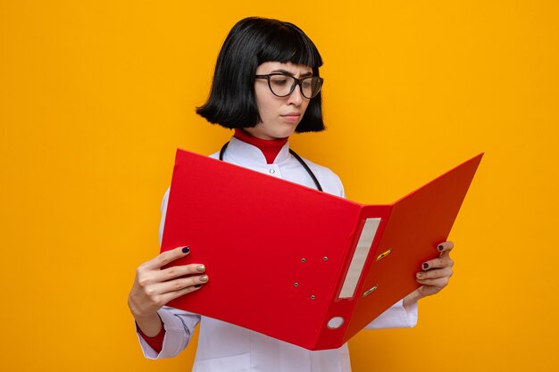 Mujer bastante caucásica joven disgustada con gafas en uniforme médico con estetoscopio mirando la carpeta de archivos