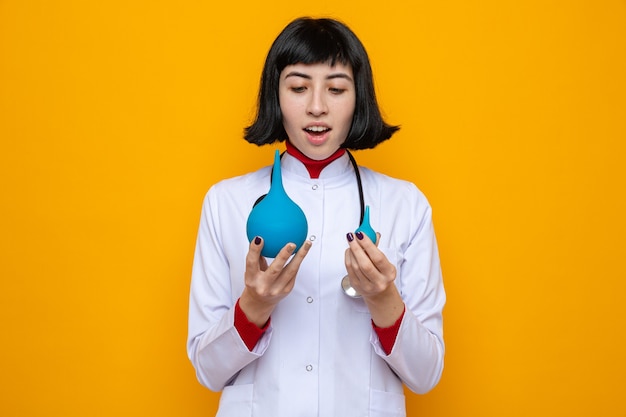 Mujer bastante caucásica joven sorprendida en uniforme del doctor con el estetoscopio sosteniendo y mirando enemas