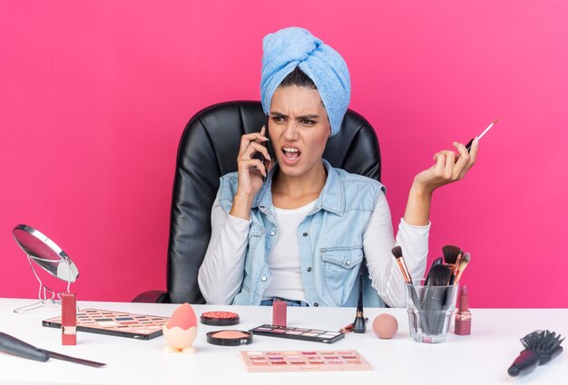 Mujer bastante caucásica molesta con el pelo envuelto en una toalla sentado en la mesa con herramientas de maquillaje hablando por teléfono y sosteniendo el brillo de labios aislado en la pared rosa con espacio de copia