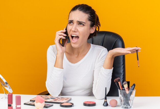 Mujer bastante caucásica molesta sentada en la mesa con herramientas de maquillaje gritando a alguien en el teléfono y sosteniendo el pincel de maquillaje aislado en la pared naranja con espacio de copia