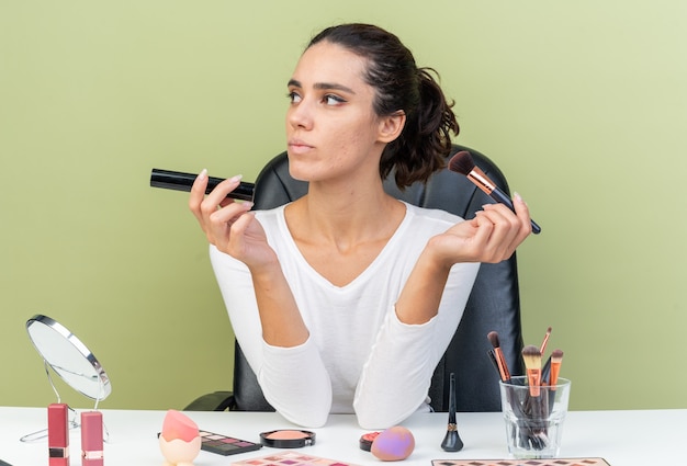 Mujer bastante caucásica segura que se sienta en la mesa con las herramientas del maquillaje que sostienen el cepillo del maquillaje y que mira el lado aislado en la pared verde oliva con el espacio de la copia