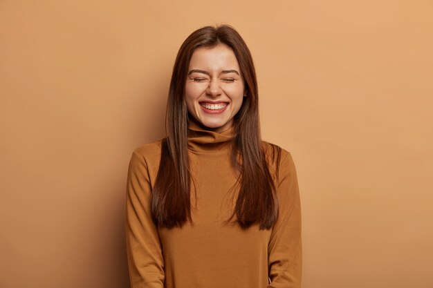 Una mujer bastante divertida tiene el pelo largo y oscuro, se ríe de alegría, viste un suéter de cuello marrón, sonríe con satisfacción