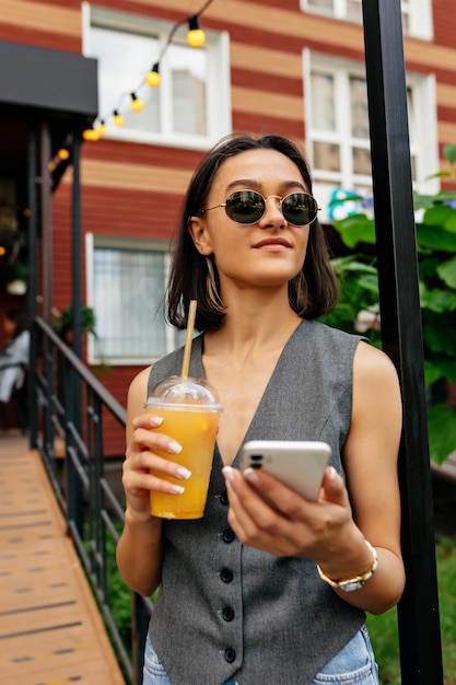 Una mujer bastante feliz con un peinado corto y oscuro con gafas de sol sostiene una bebida de verano y un teléfono inteligente y mira a un lado