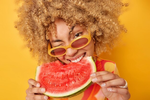 Una mujer bastante guapa con pelo rizado y tupido que muerde una sandía jugosa y apetitosa usa gafas de sol de moda disfruta comiendo deliciosos bocadillos de verano poses de frutas favoritas contra el fondo amarillo del estudio