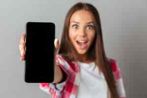 Foto gratuita mujer bastante joven asombrada que muestra el teléfono de la pantalla en blanco, foco selectivo
