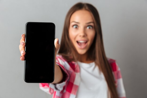 Mujer bastante joven asombrada que muestra el teléfono de la pantalla en blanco, foco selectivo