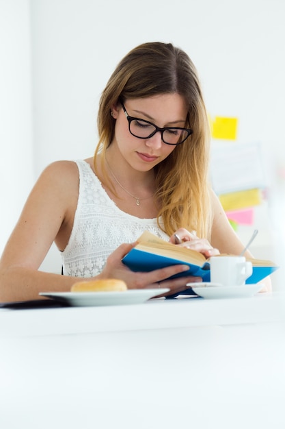 Mujer bastante joven leyendo un libro y desayunando en casa.