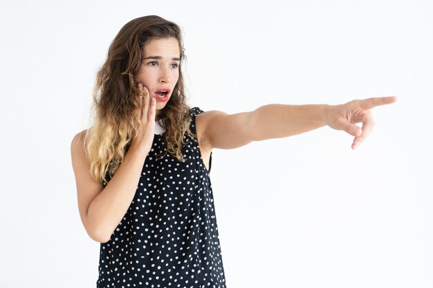 Foto gratuita mujer bastante joven preocupada que señala lejos y que toca la mejilla. concepto de incidente