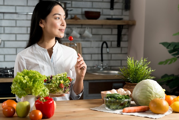 Mujer bastante joven que come el tomate de cereza rojo que sostiene el cuenco de ensalada mezclada