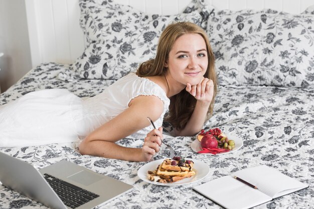 Mujer bastante joven que miente en cama con el desayuno; libro y laptop