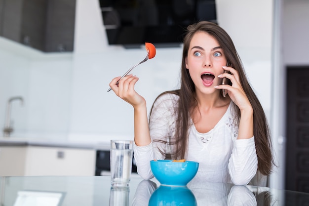 Mujer bastante joven que usa su teléfono móvil mientras que come la ensalada en la cocina en casa.