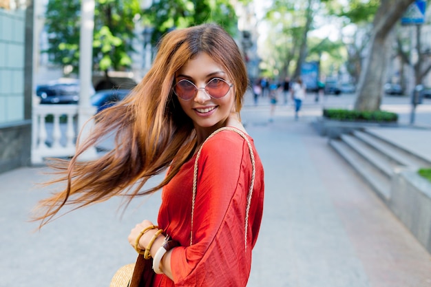 Mujer bastante joven romántica que mira sobre hombro mientras que presenta en la ciudad europea soleada del verano. Vestido coral brillante.