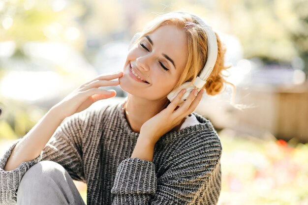 Mujer bastante joven en suéter gris disfruta de la canción mientras se sienta en los auriculares en la plaza de la ciudad