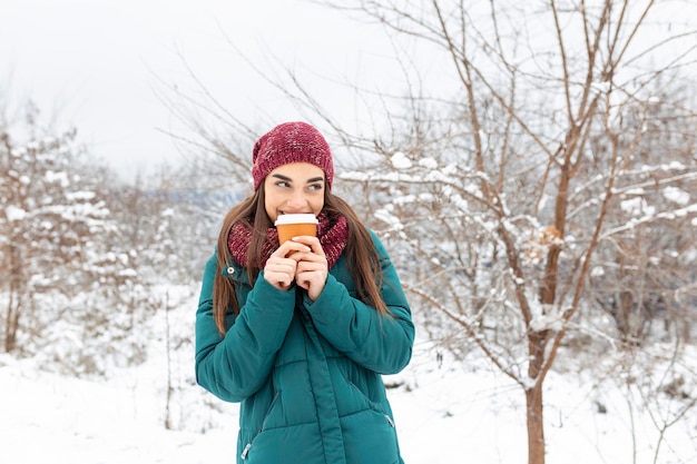 Mujer bastante joven en traje de invierno sosteniendo una taza desechable llena de café o té caliente Chica sosteniendo una taza de bebida caliente en sus manos y camina al aire libre en invierno